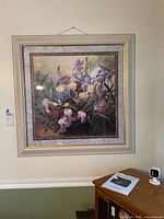 Square floral painting with irises in a white and gold frame hanging on a wall above a wooden cabinet.