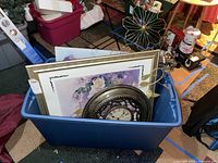 Bin containing framed floral wall art and a round metal-framed clock among other items