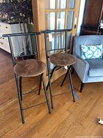 Front and partial side view of a pair of black metal folding barstools with brown cushioned round seats placed indoors on wooden floor.