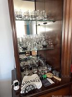 Full view of the wooden display cabinet with multiple shelves holding an assortment of rocks glasses, shot glasses, and champagne flutes. The bottom shelf holds various bar tools and accessories including a Jack Daniel's bar towel.