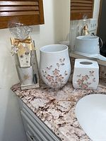 White ceramic waste bin and tissue box cover with pink floral pattern displayed on a bathroom counter next to wrapped bath soap and mirrored wall.