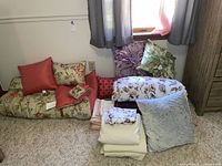 Three bed linen sets including folded sheets, pillowcases, and accent pillows in floral and bird prints with coral, lavender, blue, and green colors.