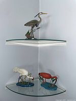 Three bird figurines displayed on corner glass shelves: one gray heron on the top shelf, two cranes (one white, one red) on the lower shelf.