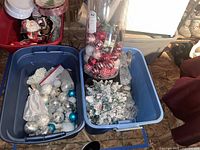 Photo shows blue storage bins with mixed Christmas ball ornaments in silver, white, teal, and some red in a separate container, alongside a small flocked tabletop Christmas tree and what appears to be lanterns and other decorations.