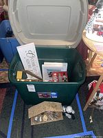Open green storage bin showing multiple Christmas decor items including framed art, toy soldiers, and white snowman figure beside nativity scene stable figurines arranged in front.