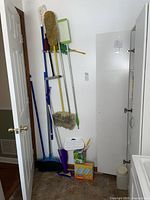 Photo showing multiple brooms, dust mop, and cleaning tools hung on a rack near a cabinet with some floor items including a Swiffer mop and cleaning products.