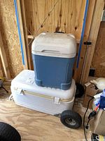 Two Igloo coolers stacked, larger white cooler on bottom, smaller blue cooler on top. Both have pull handles and wheels, visible wear and dirt.