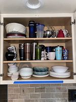 Kitchen cabinet with Corelle dishes including bowls, plates, some decorated, travel mugs and various drinkware items