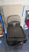 Photo showing beige folding table with rust spots leaning against wall, two gray folding chairs stacked in front, and dark brown plastic garbage can with bag inside.