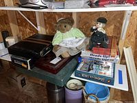 Shelf with a collection of board games including Middle River-opoly, Heads Up!, Family Fuel, Backgammon, and Pictionary. On top are two dolls: a teddy bear in a green and white dress and a smaller child doll music box.