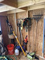 Wide shot of garden tools hanging on the wall with bucket and sprayer on floor, showing Fiskars tree cutter and Black and Decker hedge trimmer.