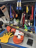 Photo showing the variety of hand tools including saw, crowbar, pipe wrench, hammer, pliers, gloves, and level on a metal workbench with extension cords coiled below.