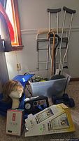 Photo showing walker, crutches, canes, wigs on Styrofoam head, compression system device, hand weights and other medical accessories stacked on carpet floor near window