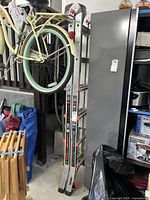 Vertical multi-position aluminum ladder folded and stored upright in garage area, near bicycle and storage cabinet.