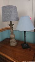 Two table lamps on wooden surface, one with wood turned base and gray shade, the other with metal square base and light blue shade