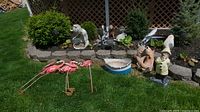 Wide view of lawn and garden bed with most items grouped together including metal flamingos, birdbath, turtles, metal dog and cat, stone sailor and fish.