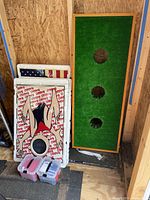 Two stacked cornhole boards with Baggo logo and cartoon figure design by wall inside wooden shed, accompanied by plastic containers with bean bags.