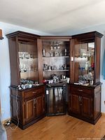 Full front view of the lighted corner entertainent cabinet showing glass shelves, lighting, mini fridge, and base cabinets with countertop surfaces and doors.