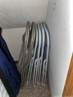 Six metal folding chairs folded and leaned against a wall in a corner, showing two color variations of frames: grey and white with blue speckles