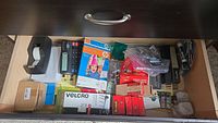 Drawer with assorted office supplies including tape dispenser, calculator, photo paper, batteries, Velcro strips, stapler, notebooks, and small office items.