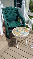 Four stacked green outdoor chairs with woven vinyl strap seats and backs on a deck with a small folding table with a colorful floral top beside them.
