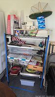 Full view of metal shelf with all party hosting supplies: cups, napkins, aluminum foil, bags, flags, citronella holders, trays, plastic bins, grill tools.