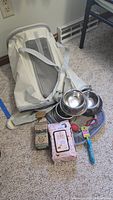 Photo showing white pet carrier with mesh ventilation, straps, three stainless steel pet bowls, cat toy, new cat collar in package, unopened pet wipes, and opened poop bags box arranged on a carpeted floor.