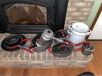 Wide view showing a mix of cookware including Rachel Ray red-handled non-stick pans and saucepans, white enamel pots and a metal kettle placed on a brick hearth.