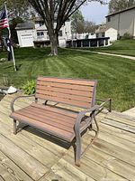 Rocker bench with slatted wooden seat and backrest, attached curved metal armrest, placed on wooden dock near water. Rust visible on metal parts.