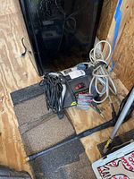 Side and front view of Ryobi electric pressure washer unit showing power cable, soap hose, and spray wand attachment placed on wooden floor.