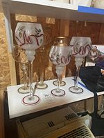 Photo showing 5 candle holders on white shelf, 3 clear glass with red and gold holly designs, 2 amber glass with gold leaf designs