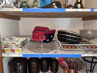 Shelf showing floral ceramic rectangular bowls, American themed ceramic bowl with a red kitchen towel inside, black wicker bowls in white wire tray, clear glass dessert tray, and brown canisters below.