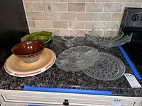 View of ceramic and assorted clear glass serving dishes on granite countertop. Shows two green leaf-shaped glass dishes, a clear glass oval dish, a divided clear glass round plate, multiple clear glass plates with oval and round shapes, and ceramic bowl and plate in front.