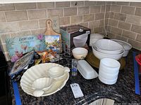 Photo showing various white ceramic serving dishes including a large shell-shaped dish with two smaller dishes, stackable bowls, butter dish, metal spice rack, glass sand timers, cutting boards and cocktail-themed hot mitts and trays on a granite kitchen counter.