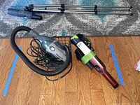 Shark and Pensar hand vacuums placed on wooden floor with attachments and a metal tripod in background.