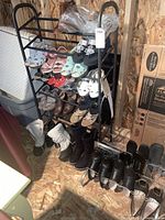 Photo showing two black metal shoe racks filled with various pairs of women's shoes including boots, sandals, and casual shoes, plus additional shoes on the floor nearby.