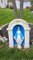 Concrete statue of Mary in white and blue positioned inside white concrete archway with blue interior and angel figure on top, situated on grass near trees and stone border.