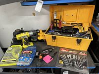 Wide shot showing yellow Ryobi drill, Worx cordless screwdriver, Ryobi and Skil drill bit sets, and yellow toolbox with hand tools and small hardware.