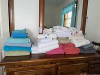 Wide shot showing a full assortment of towels and hand towels folded and stacked on a wooden chest, with variety of colors and embroidered designs.