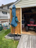 Photo of closed teal patio umbrella standing upright on grass next to shed door.