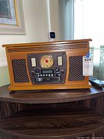 Front view of the Victrola entertainment center showing the wooden console with the analog radio dial, speaker grille on left, control buttons, and a remote control beside it.