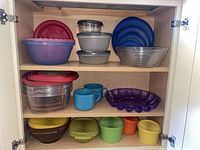 Cabinet shelves filled with various plastic containers, bowls and mugs in multiple colors and sizes including red, blue, purple, grey, clear.