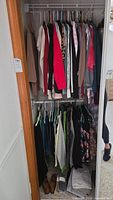 Closet image showing a variety of women's shirts, sweaters, pants, and scarves on hangers, with some shoes on the floor.