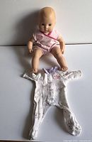 American Girl baby doll wearing pink onesie sitting next to white footed sleeper with purple designs