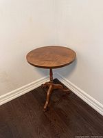 View of antique three legged round wooden table with visible wood grain, placed near a white wall and dark hardwood floor.