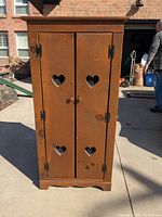 Front view of wooden cabinet with two doors, each door having two heart shaped cutouts with mesh backing. Black metal hinges and round wooden knobs visible. Cabinet stands on four feet with slight damage noted on one foot.