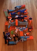 Top-down view of 8 Nerf blasters arranged on floor with multiple orange and yellow clip magazines, drum magazine, and roll of foam darts.