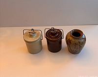Three small antique crocks and a pottery vase shown together on a white surface. Two crocks have metal handles and lids, beige and brown colors, alongside a mottled brown signed vase.