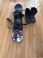 Full view of the snowboard with bindings attached and the snowboarding boots shown side by side on a hardwood floor.