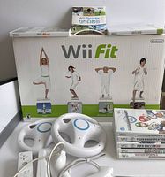 Photo of the Wii Fit balance board box, steering wheel accessories, Wii remote controllers and cables, and a clear view of stacked games.